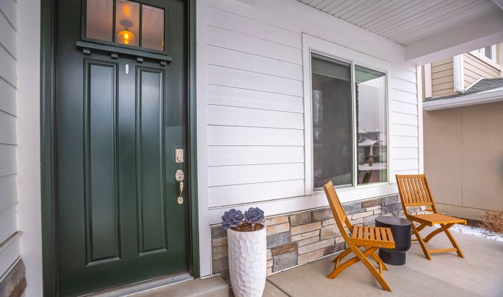 dark green door of a porch with siding and under the deck painted with white, a window, two wooden chairs and a white plant pot